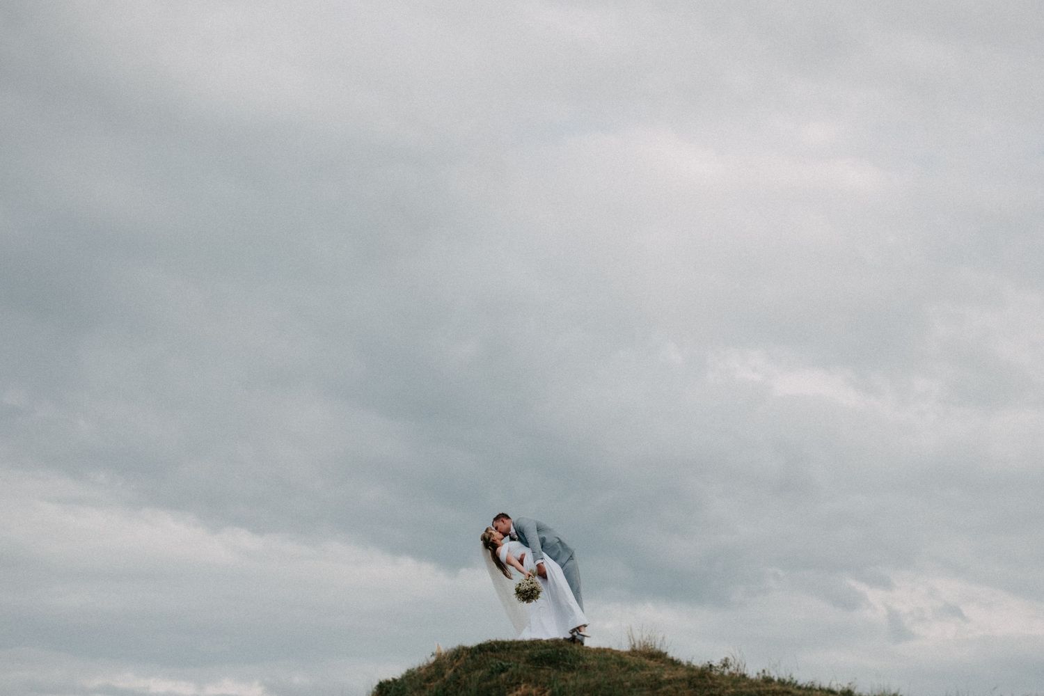 Wedding couple stands dramatically posed on hilltop against moody cloudy sky.