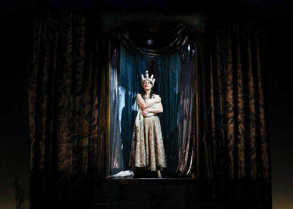 A figure in a vintage floral dress stands dramatically framed between dark curtains with moody blue lighting.