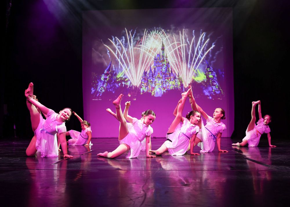 Young dancers in pink costumes perform on stage with a colorful fireworks projection backdrop during a dance recital.