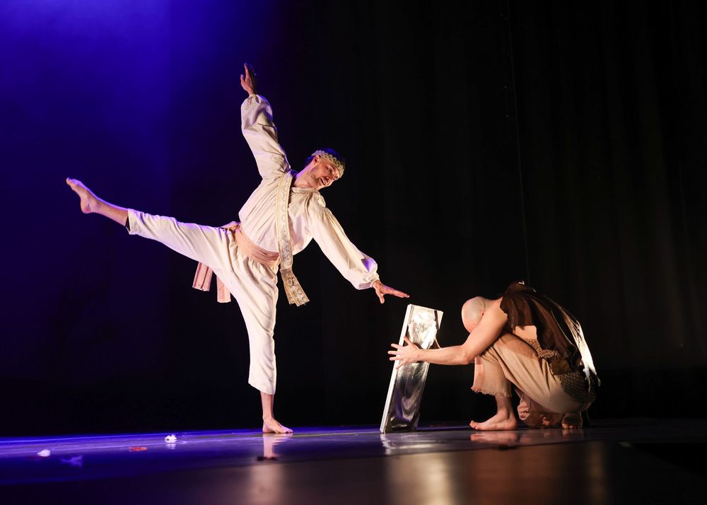 Two dancers perform an expressive contemporary dance piece on a dark stage illuminated by purple lighting.