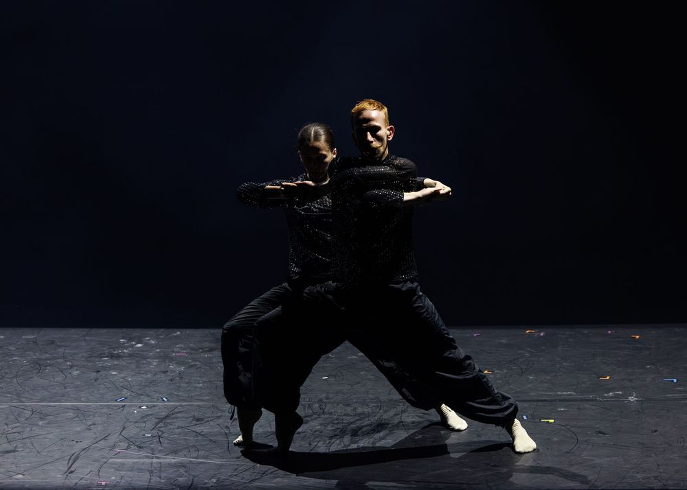 Two dancers in black costumes perform a dramatic contemporary dance routine on a dark stage.
