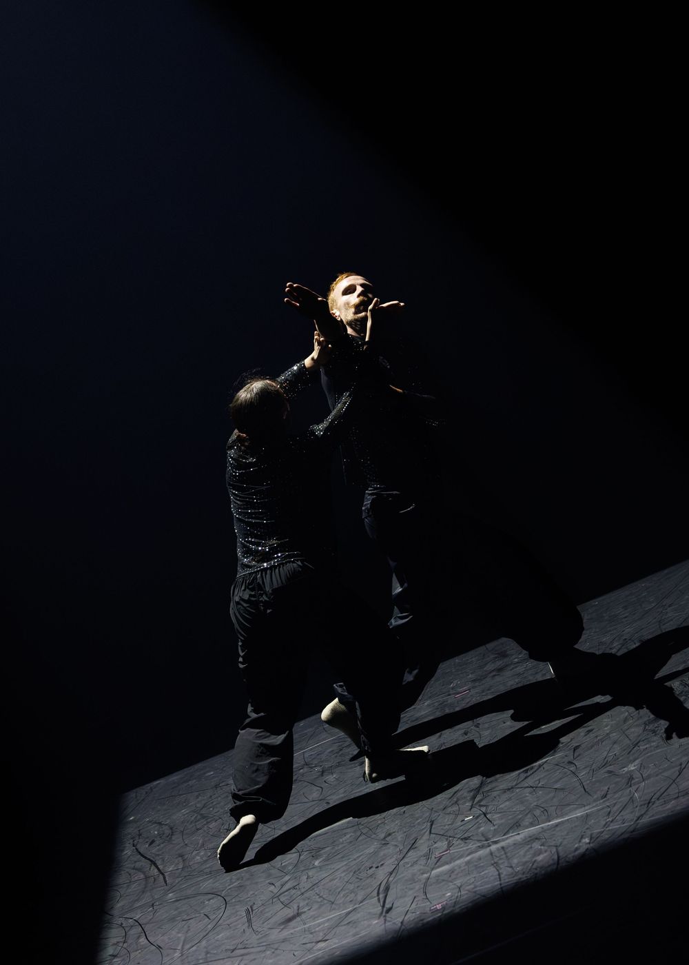 Performer illuminated by spotlight on dark stage, wearing mask, dramatic shadows cast below.