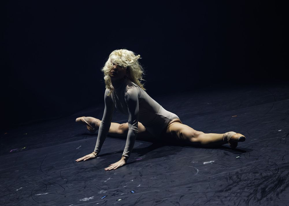 Blonde dancer performing a dramatic split pose on a dark stage floor in a gray bodysuit.