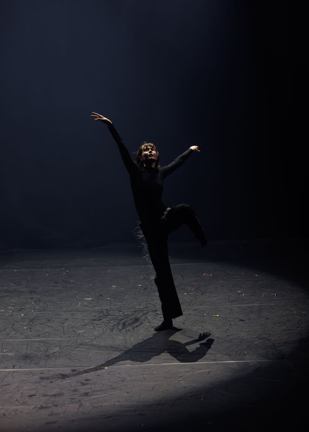 A dancer performs solo on a dark stage, dramatically lit by a spotlight, with one leg raised and arms extended.