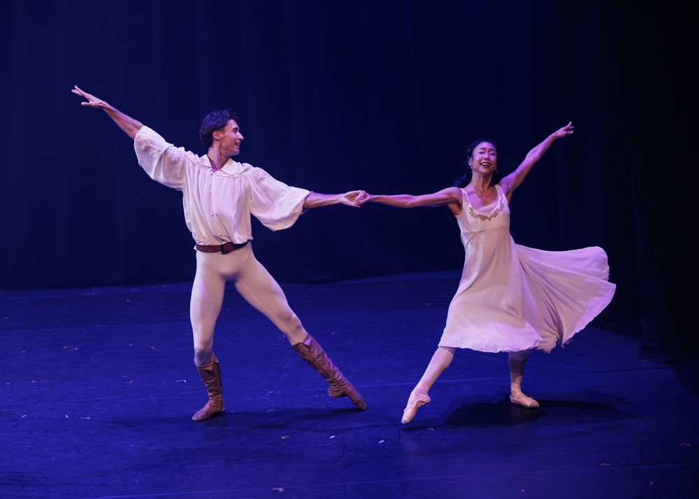 Two ballet dancers in white costumes perform an elegant pas de deux on a blue-lit stage.