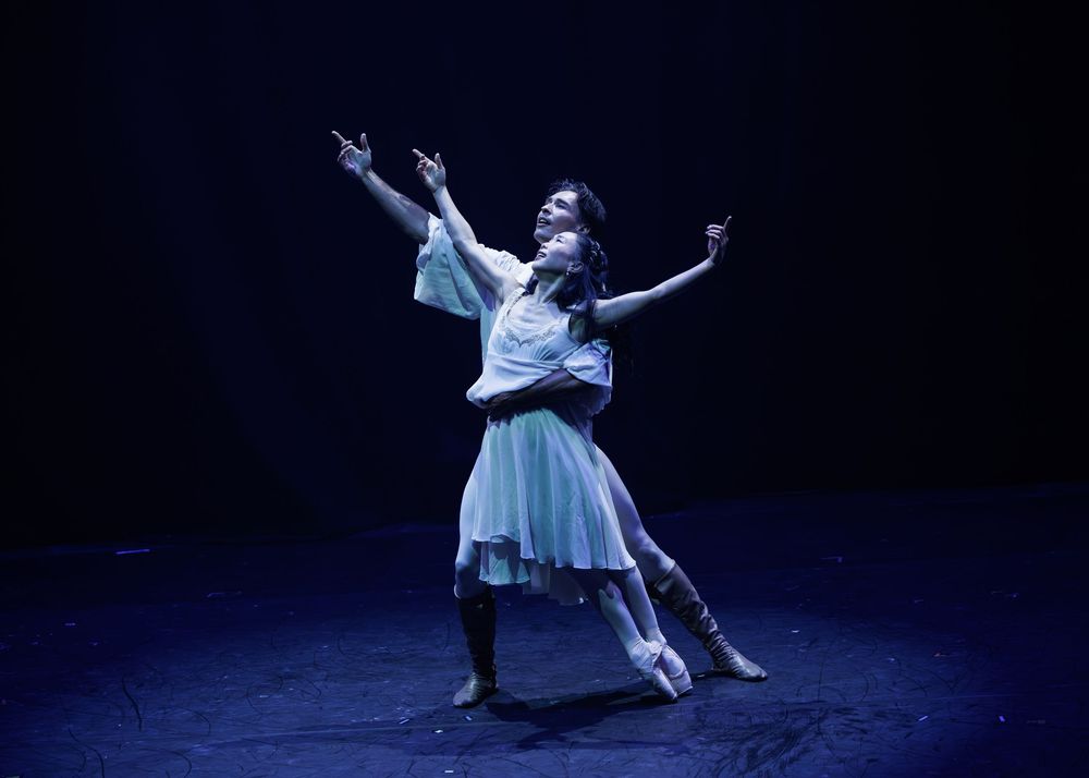 Two ballet dancers perform an expressive duet on a dimly lit stage, with graceful poses and flowing costumes.