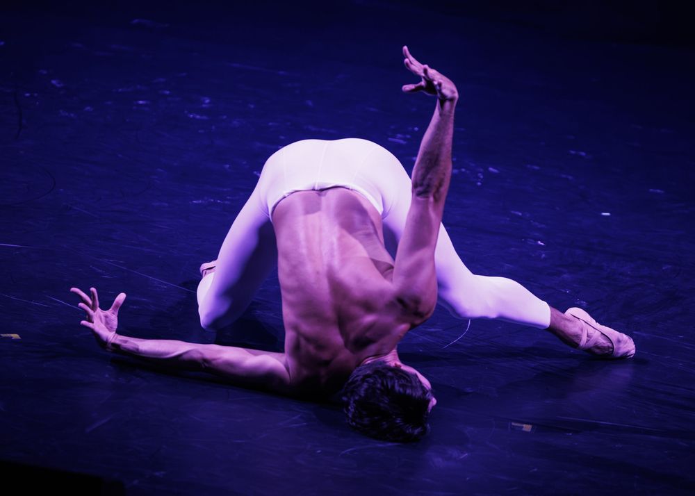 Male ballet dancer performing dramatic floor pose under purple stage lighting, showcasing strength and flexibility.