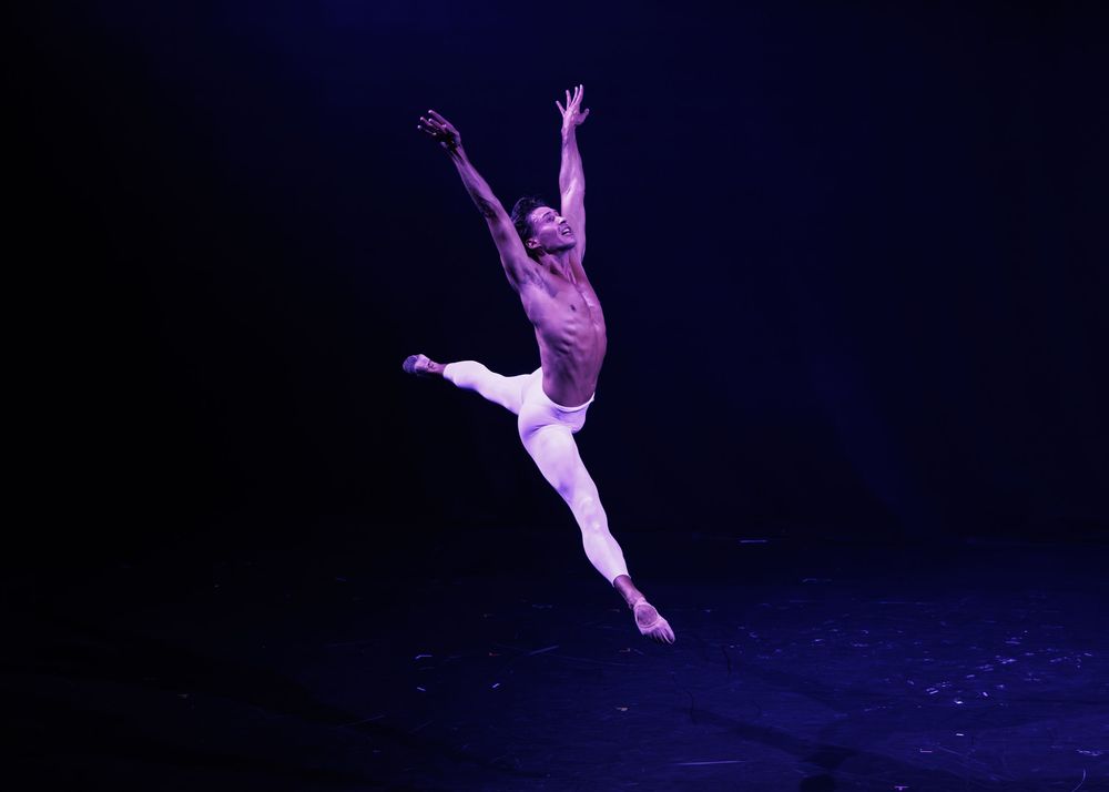 Male ballet dancer leaping gracefully on stage under purple spotlight in white tights performance.