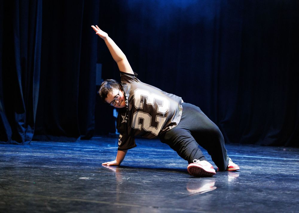 A dancer in a sports jersey performs a dramatic side lunge on a dark stage.