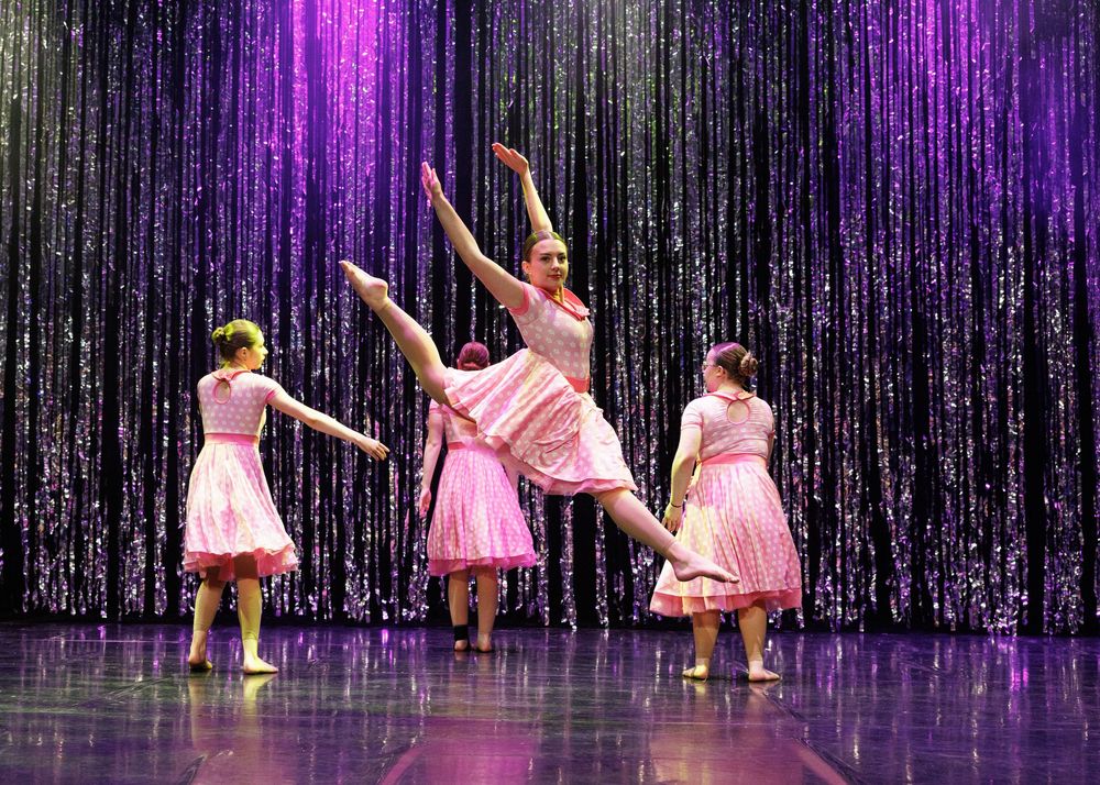 Three girls in pink dresses perform ballet on stage with a sparkling purple curtain backdrop.