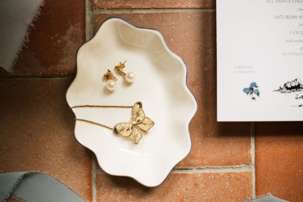 Delicate gold butterfly earrings displayed on white scalloped ceramic dishes against a rustic terracotta tile background.