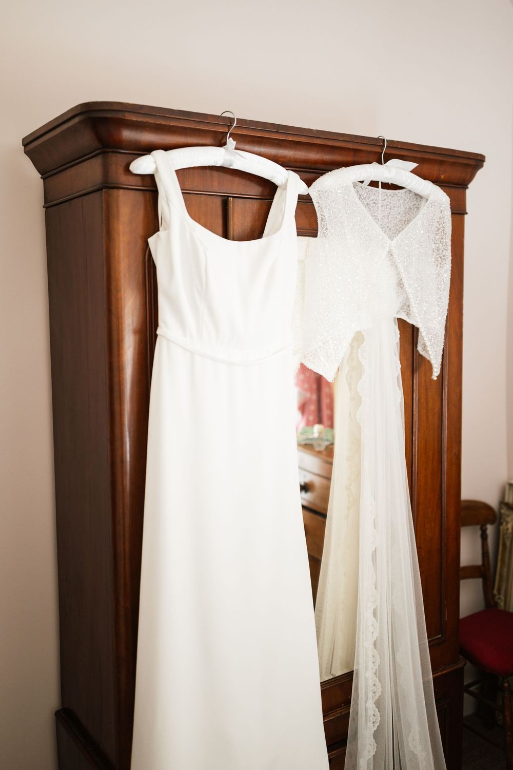 An elegant white wedding dress and lace veil hanging on wooden hangers against a dark wooden wardrobe.