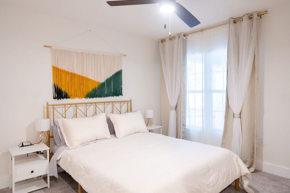 Bright bedroom with white bedding, ceiling fan, and natural light from windows.