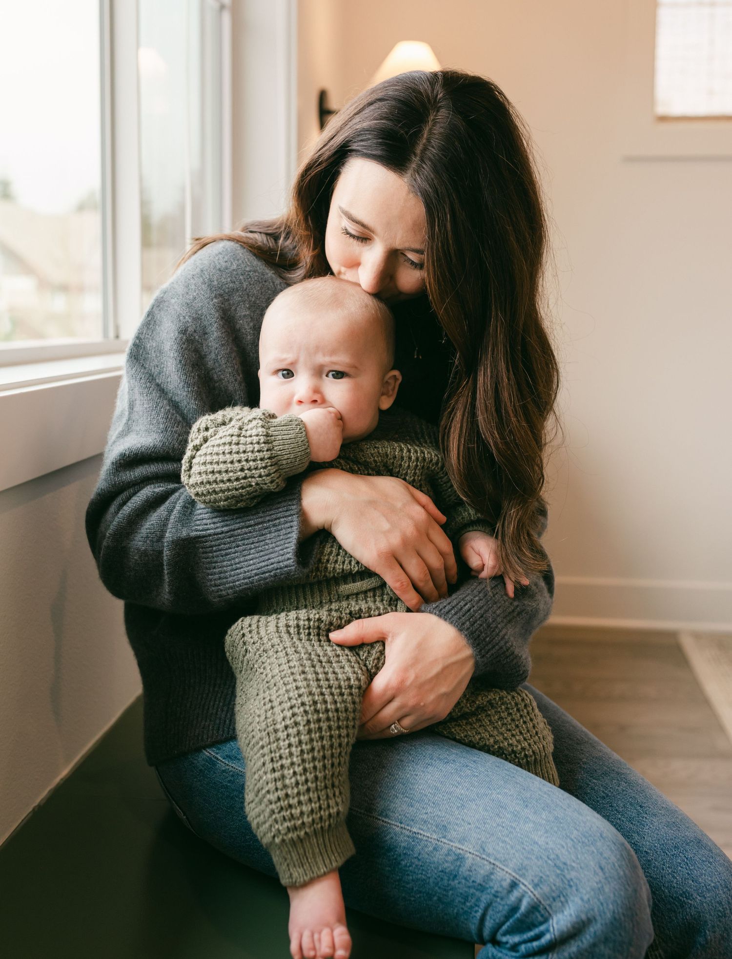 A parent in a gray sweater holds and kisses a baby wearing a knit sweater in a bright sunlit room.