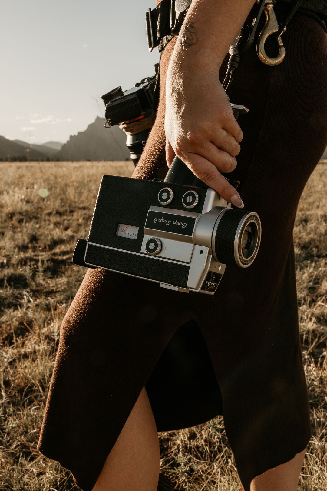 Vintage camera held against black clothing in golden sunlight during sunset or sunrise in an outdoor field setting.