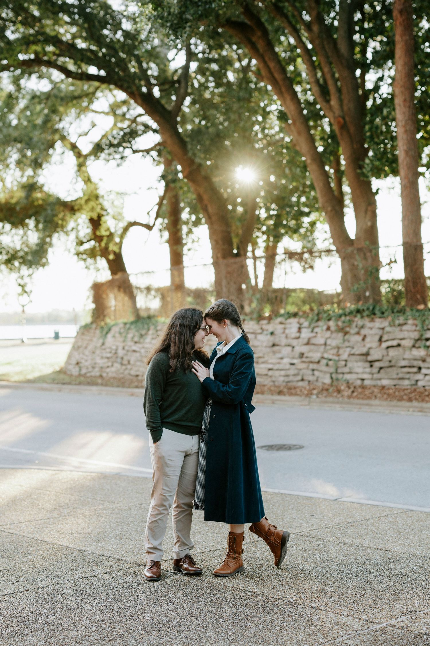 A romantic couple embraces on a sunlit path with stone walls and trees creating a dreamy backdrop.