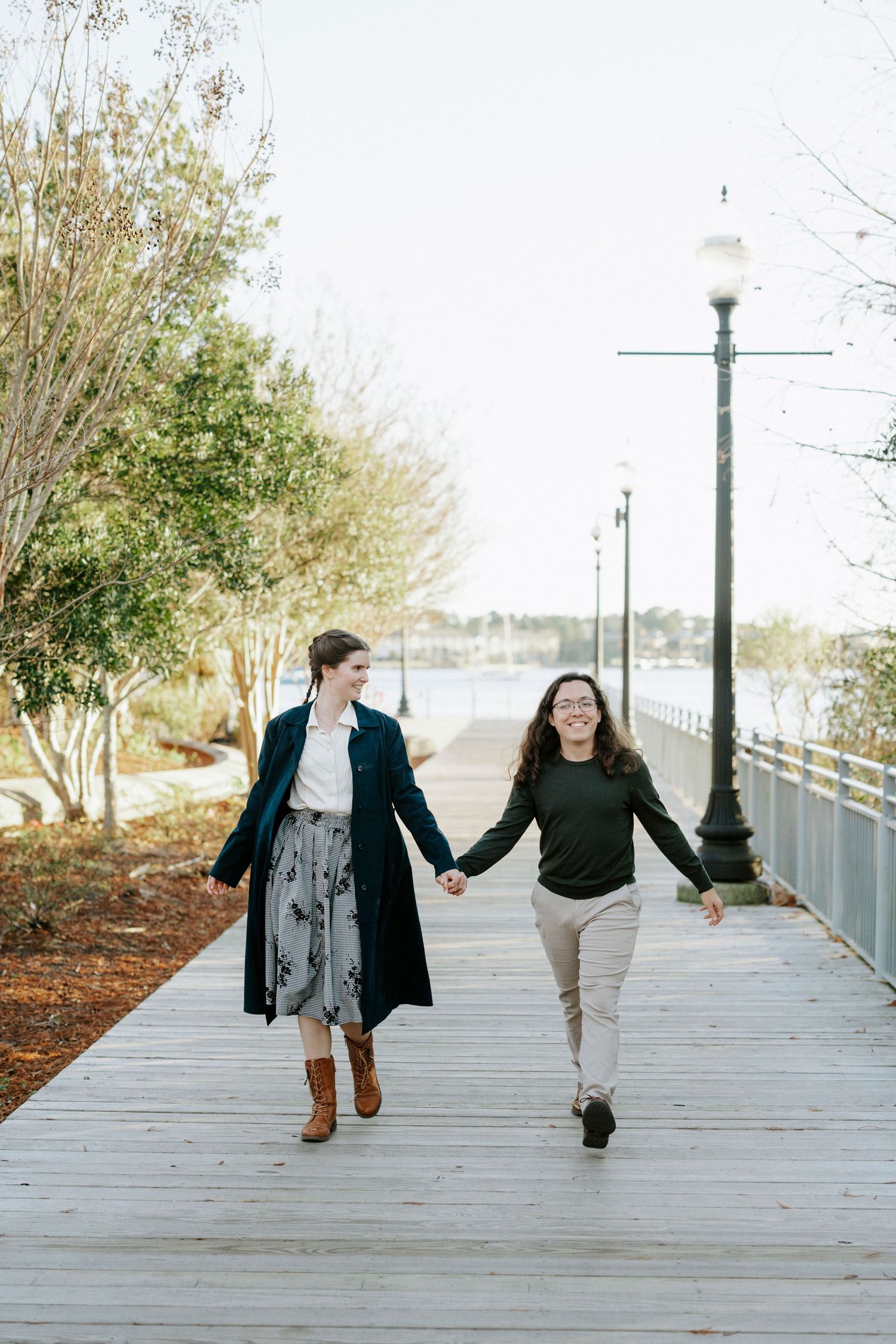 Two people playfully jump and hold hands while walking along a wooden boardwalk by the water.