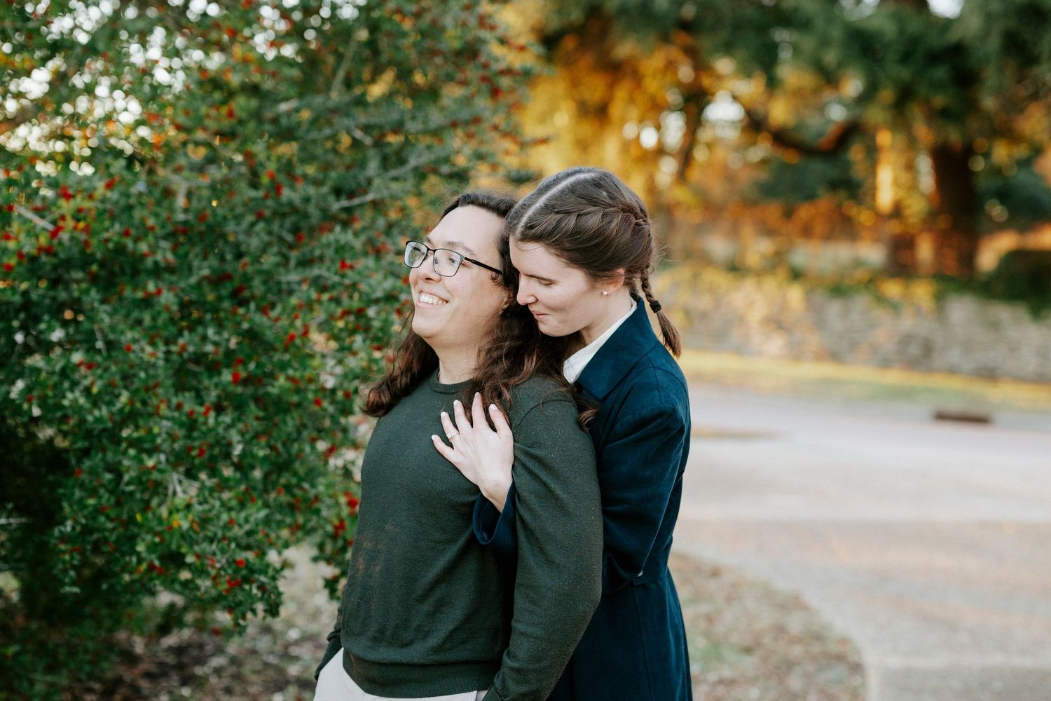 A couple embraces outdoors on a path with autumn foliage and golden sunlight in the background.
