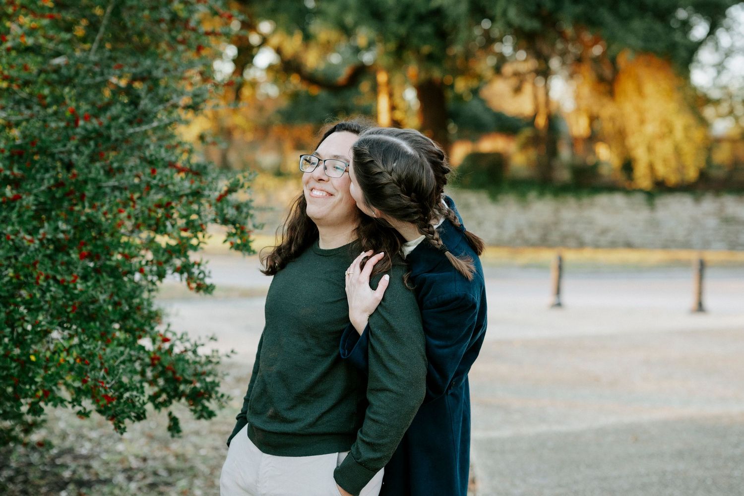 A couple in dark clothing embraces outdoors during an autumn photoshoot with golden trees in the background.