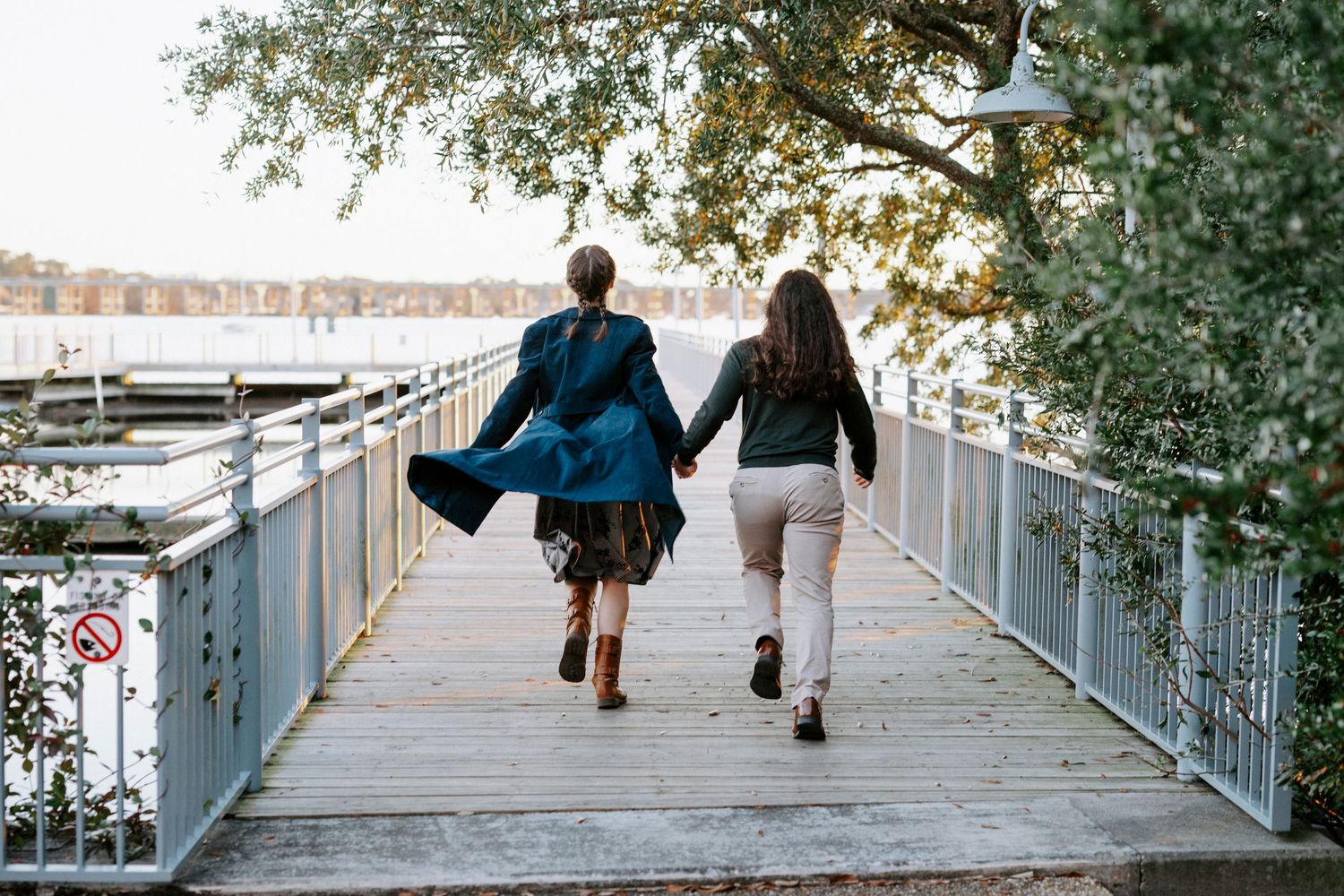 Two people walking on a waterfront boardwalk on a sunny day with boats and trees in the background.
