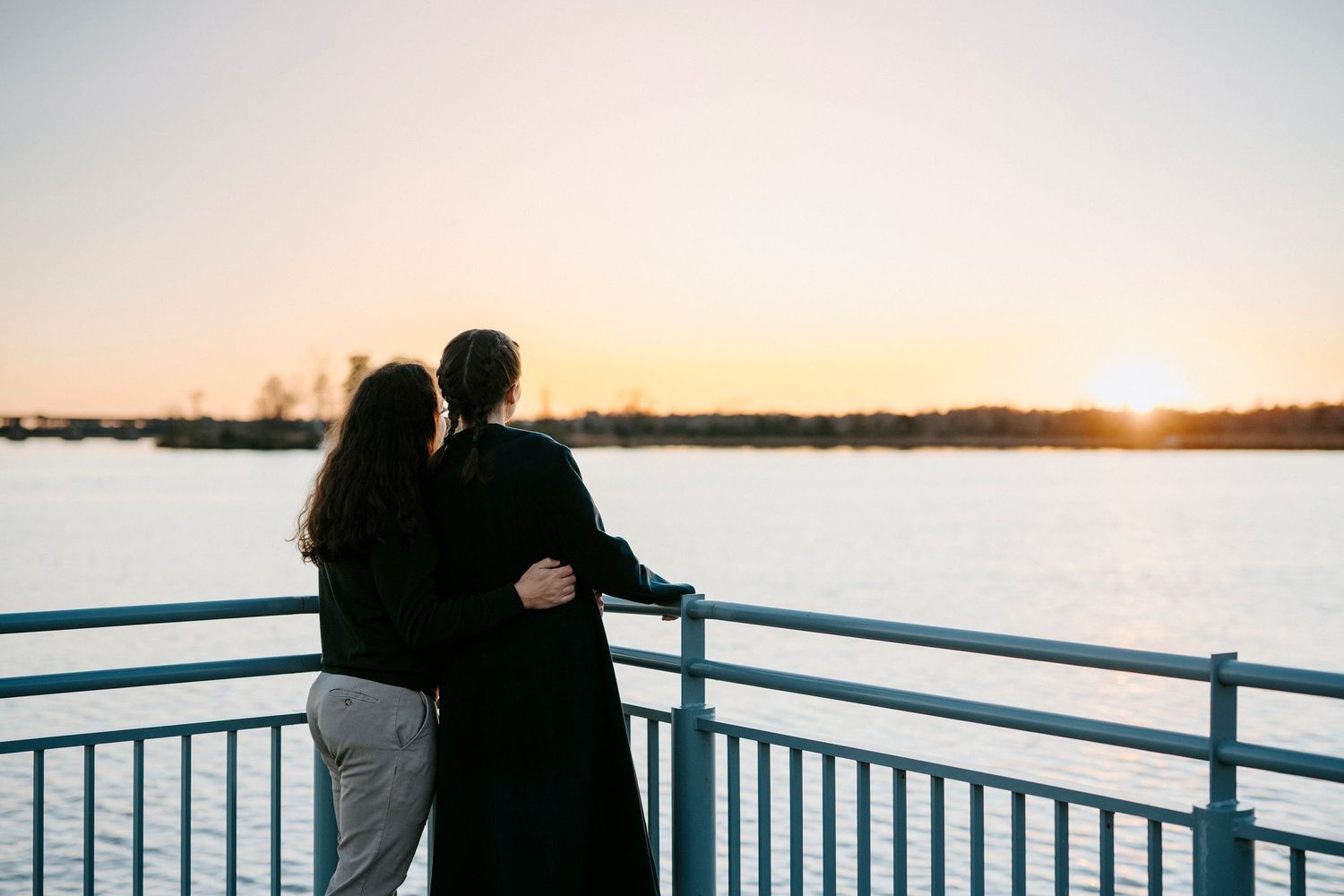 Silhouettes of a couple stand together against a waterfront sunset with railings in the foreground.