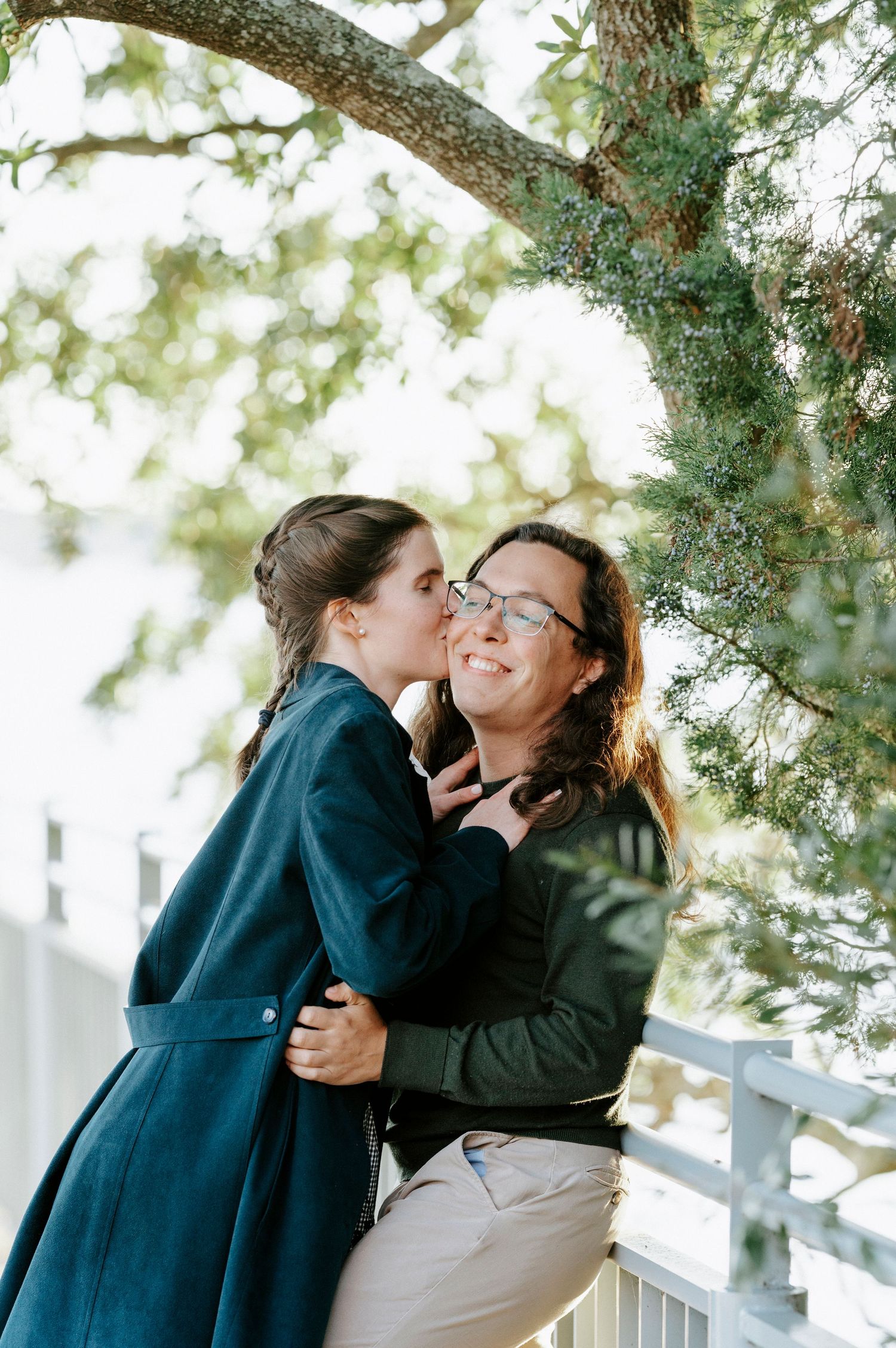 Two people share an affectionate moment under a tree branch on a bright sunny day.