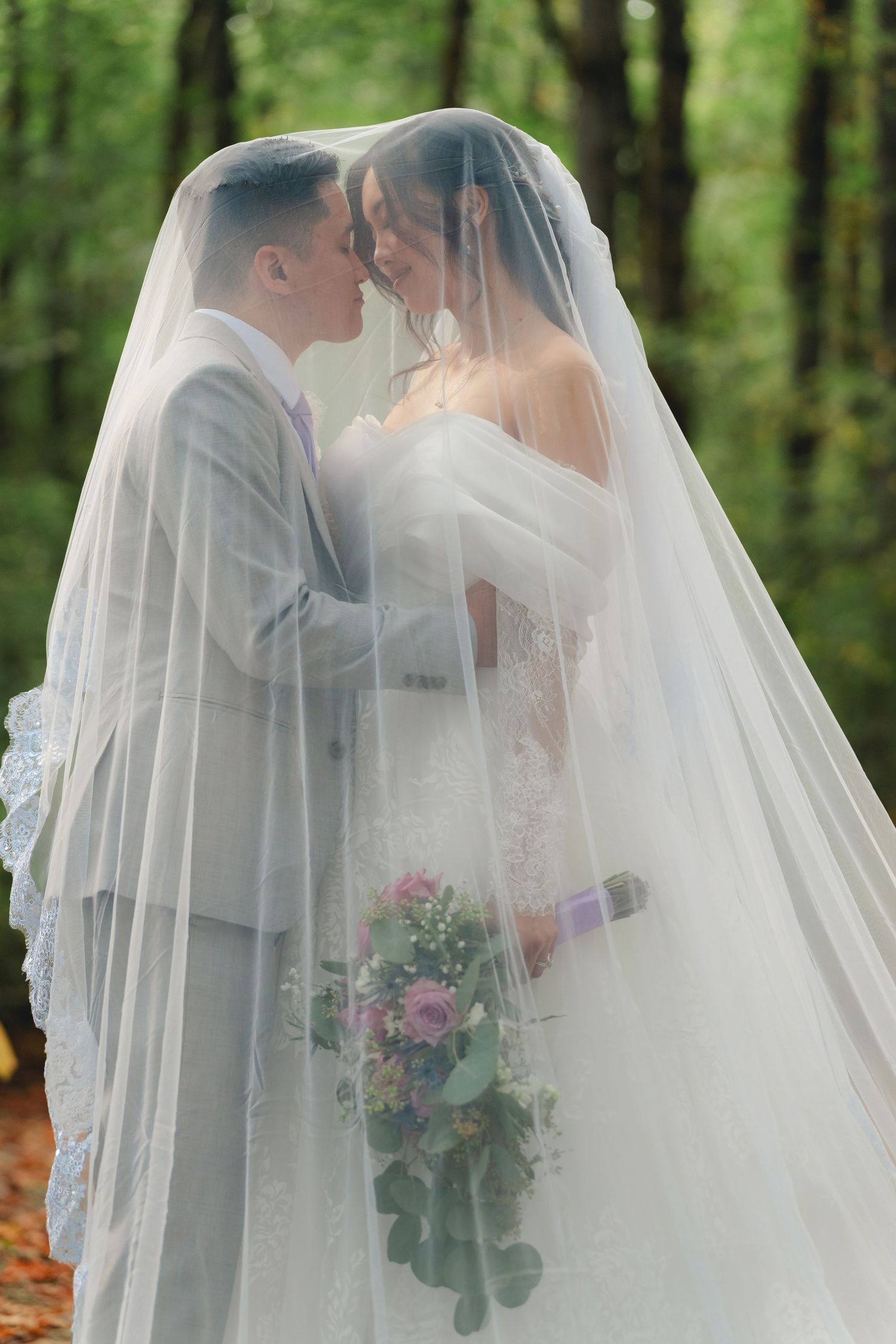 A romantic wedding couple shares a tender kiss under a flowing white veil in a lush forest setting.