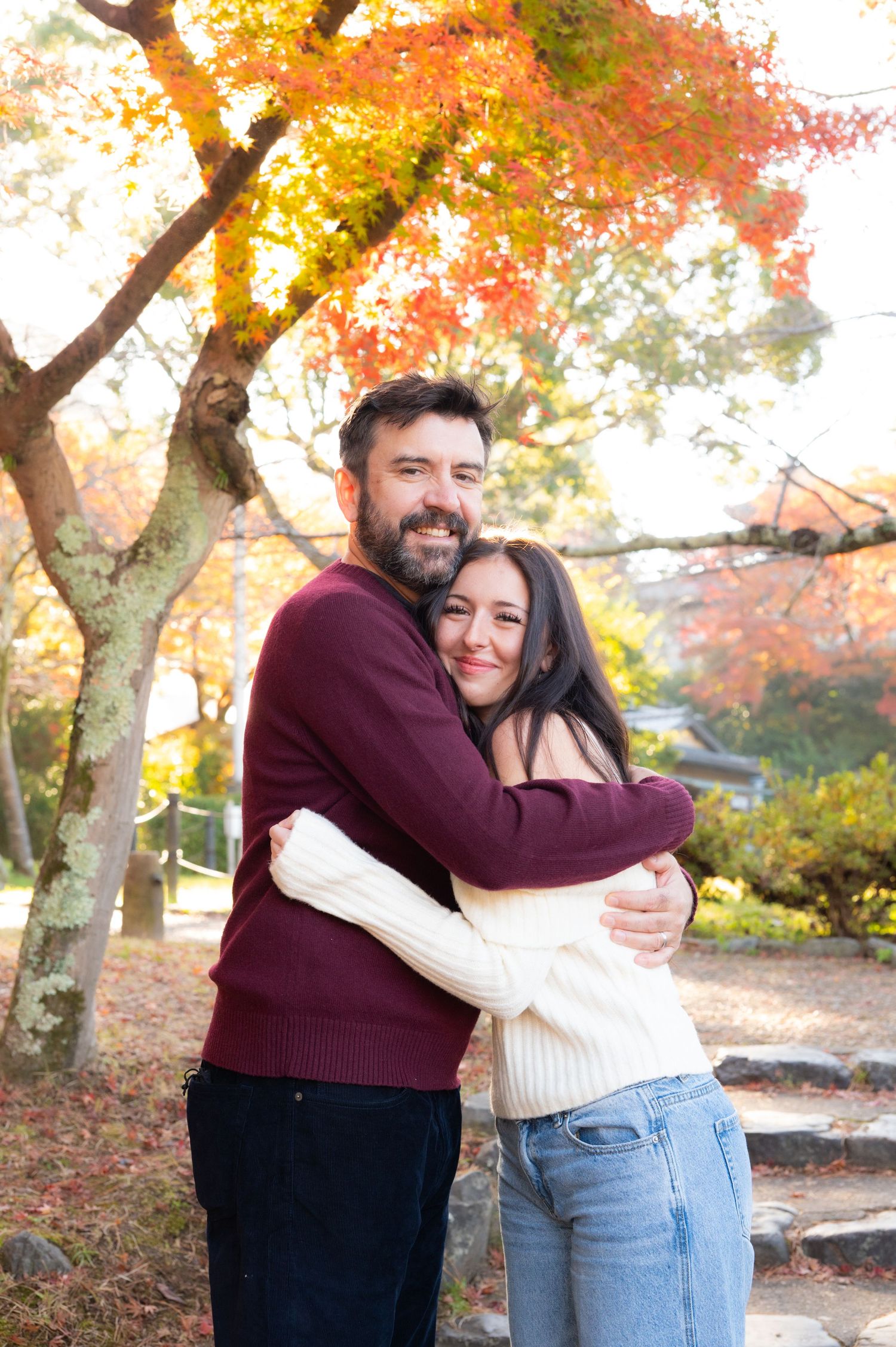 A couple hugs warmly beneath colorful fall trees in a scenic outdoor park setting.
