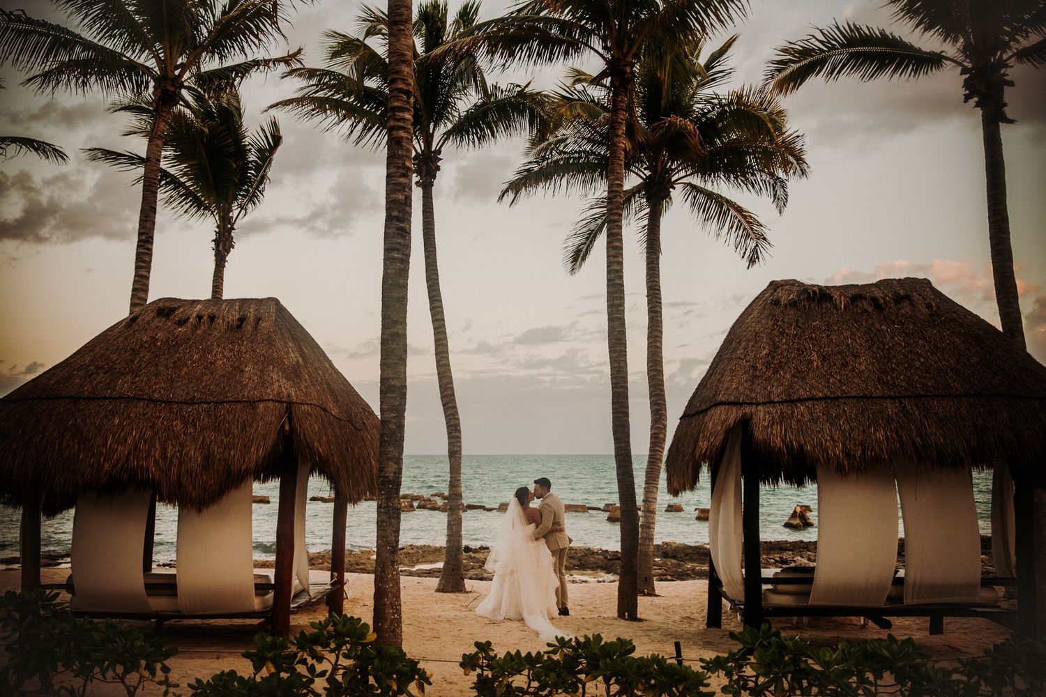 Tropical beachfront bungalows with thatched roofs line the shore among swaying palm trees at sunset. El Dorado Royale Wedding Photographer