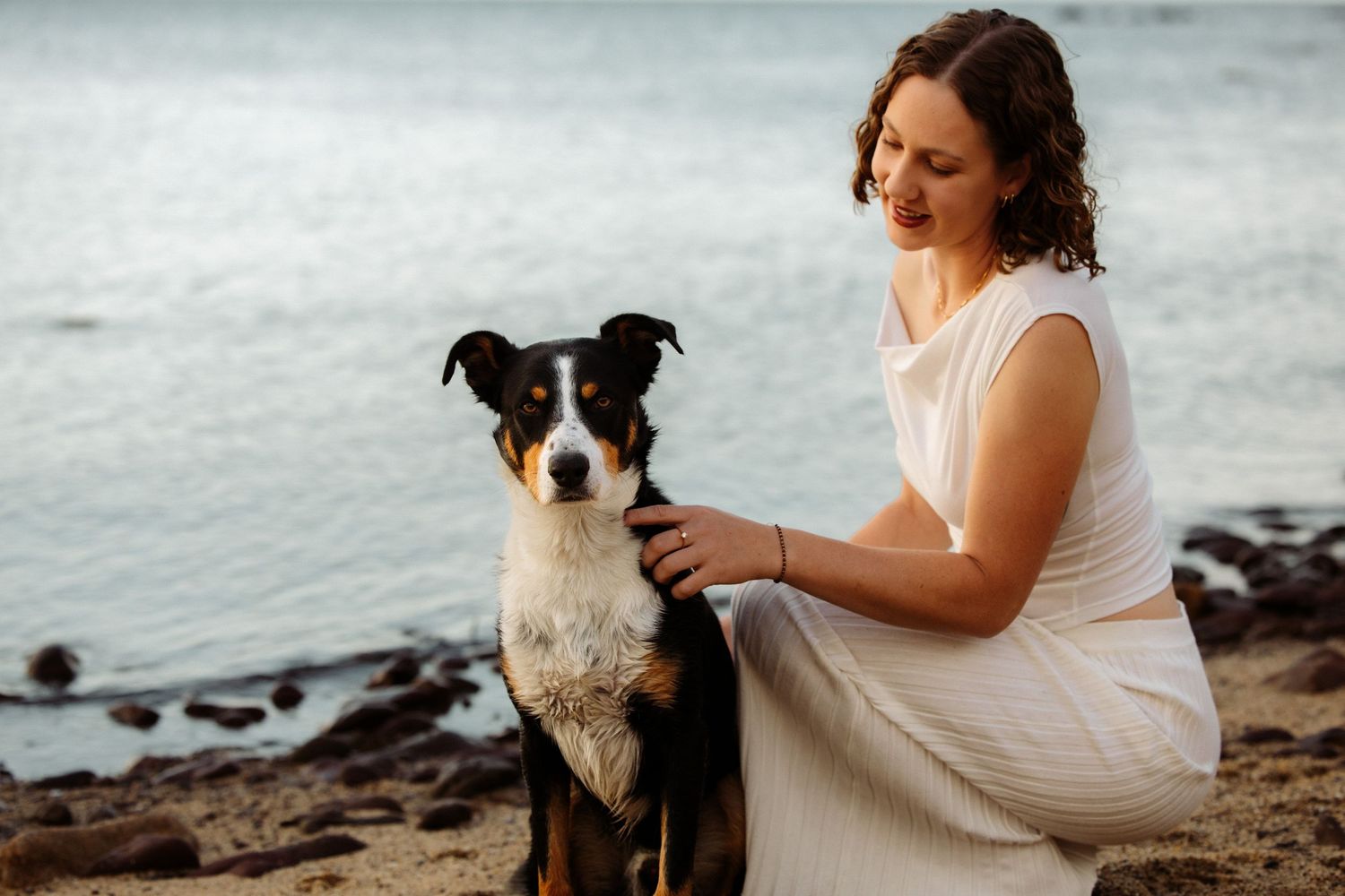 A smiling woman in a white dress sits on a rocky beach with a black and white dog near the ocean.