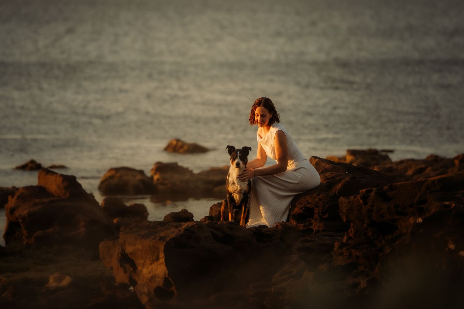 Woman in white dress sitting on rocky shore with her dog at golden hour sunset by the ocean.