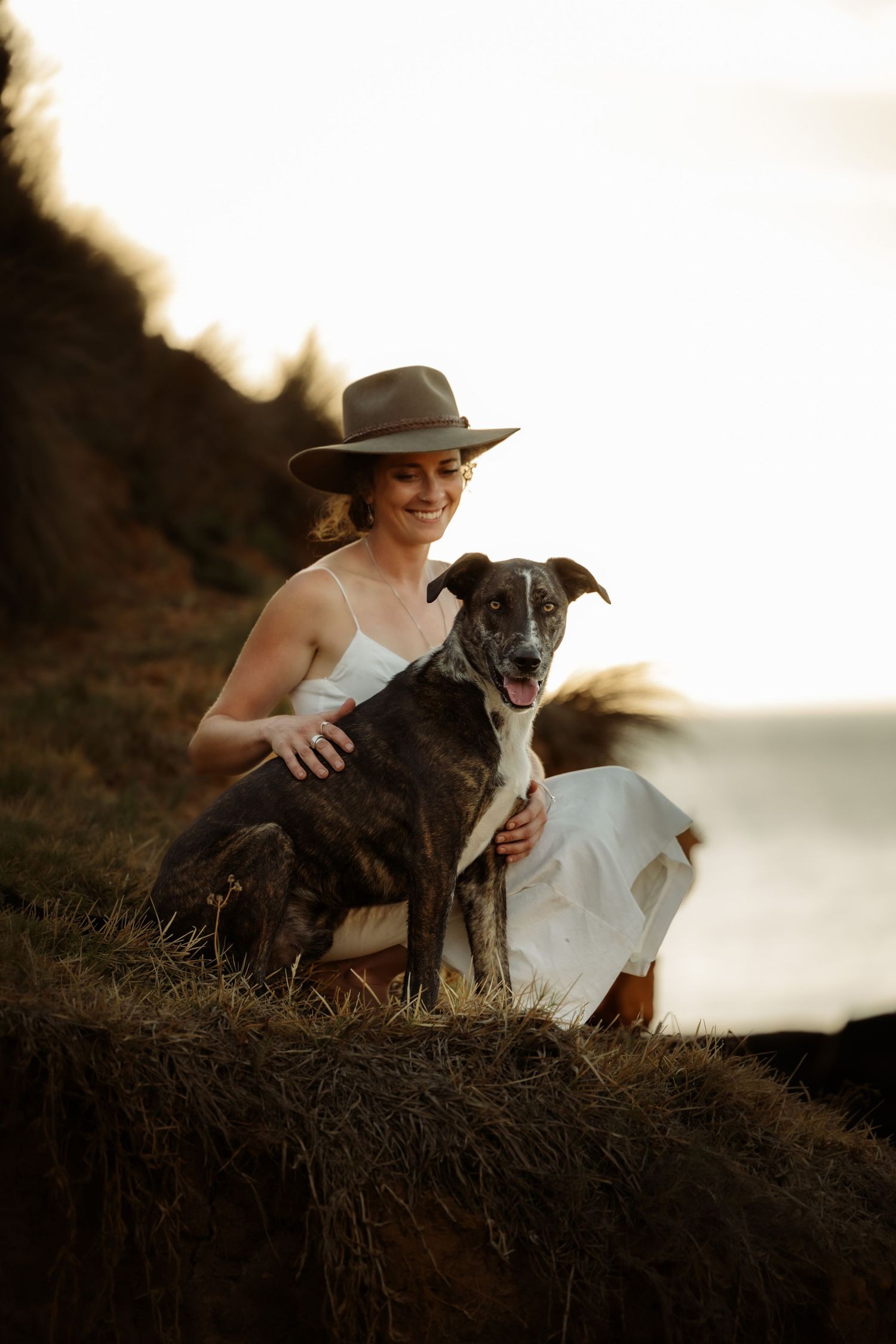 Woman in white dress and hat smiling with her dark brindle dog on a grassy cliff at golden hour.