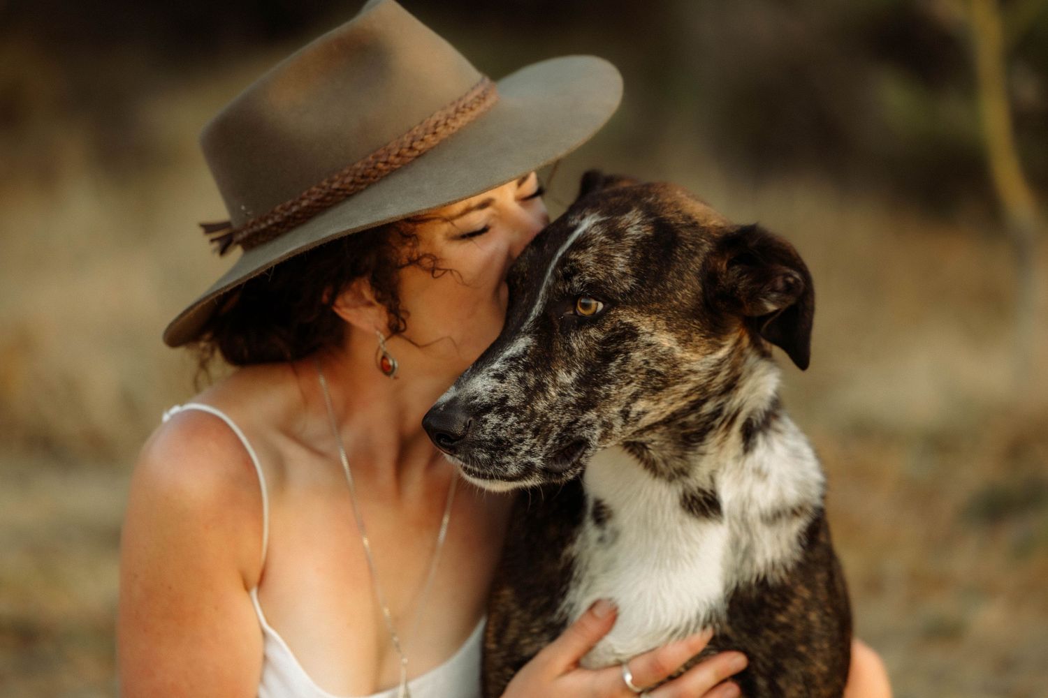 Woman in wide-brim hat kissing her brindle dog outdoors in warm natural light.