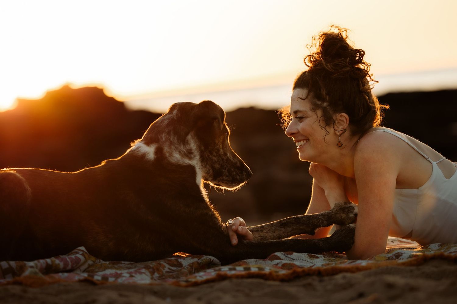 Woman smiling at her dog on a beach during golden sunset, sharing a joyful moment lying on a blanket.