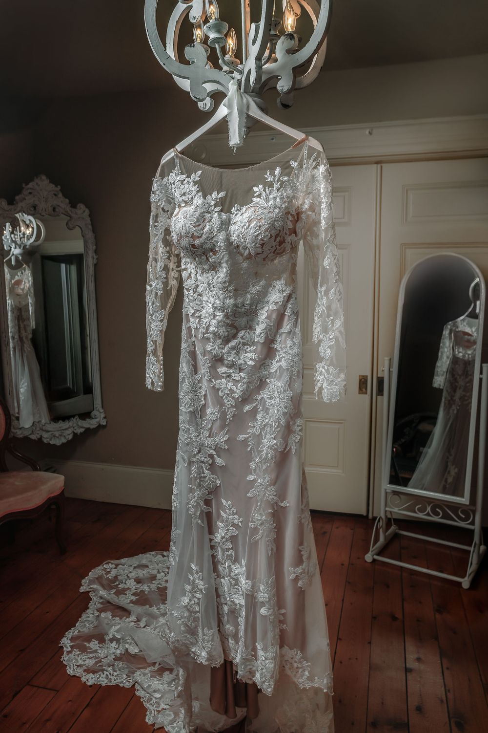 A delicate white lace wedding dress with sheer sleeves hangs from an ornate chandelier in a rustic room.
