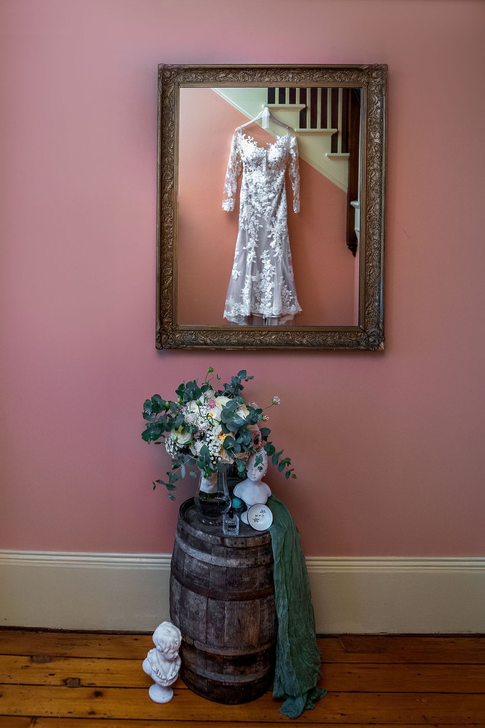 A white lace wedding dress hangs in an ornate frame above a rustic barrel with floral arrangement and green fabric.