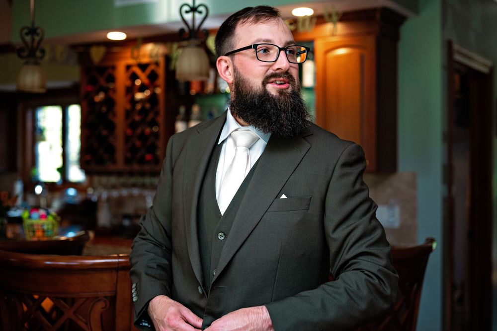 A person in a black suit and glasses stands in an elegantly lit room with ornate decor.