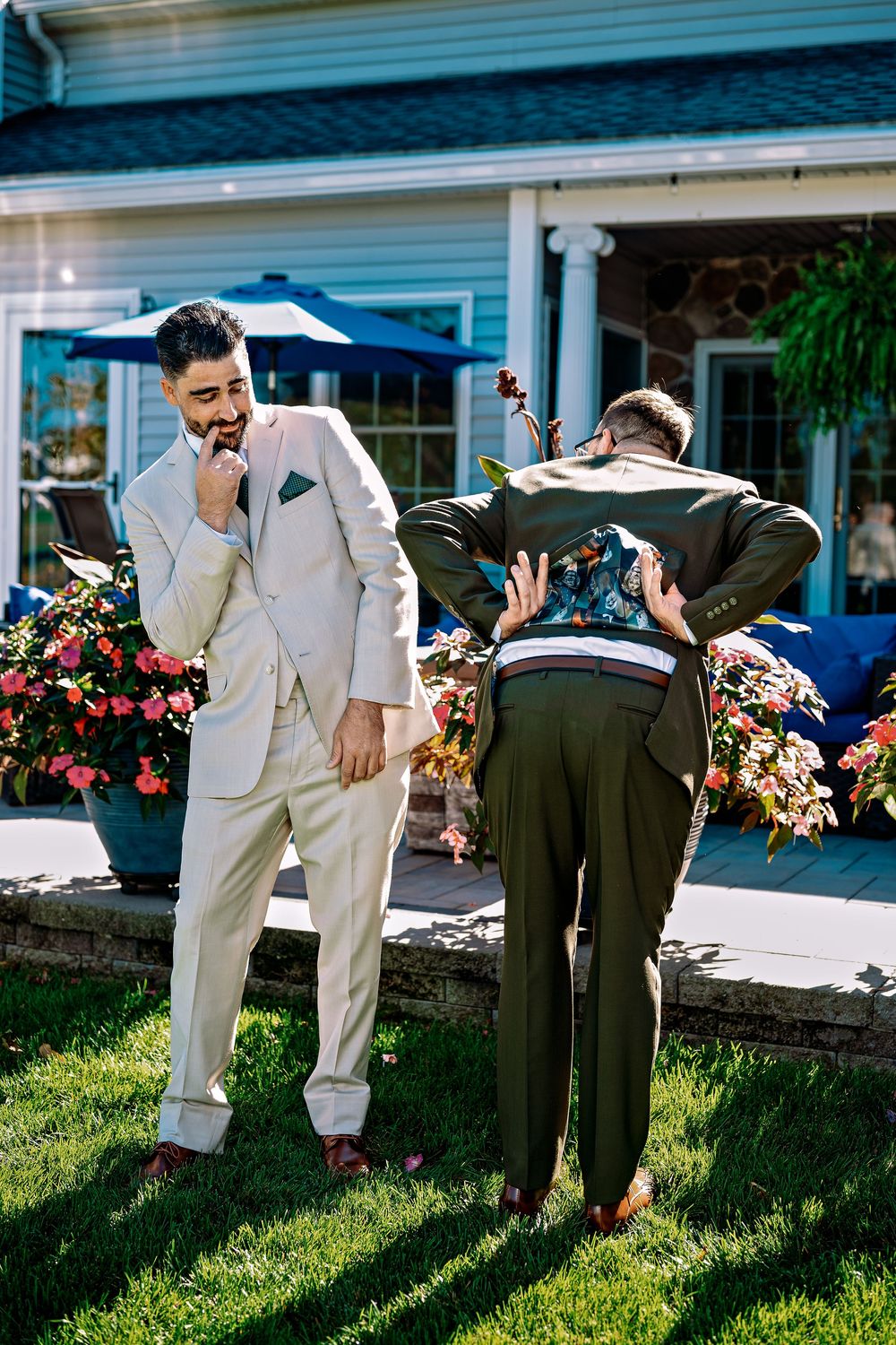 Groomsmen in light and dark suits bend over to pose playfully on a sunny lawn outside a blue house.
