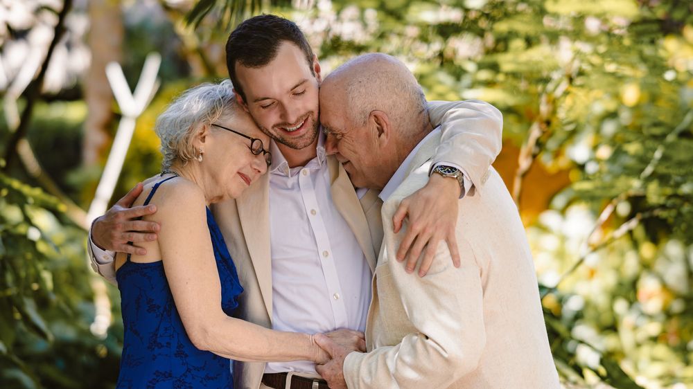Three people share an intimate moment of laughter and hugs in a sunlit garden setting.