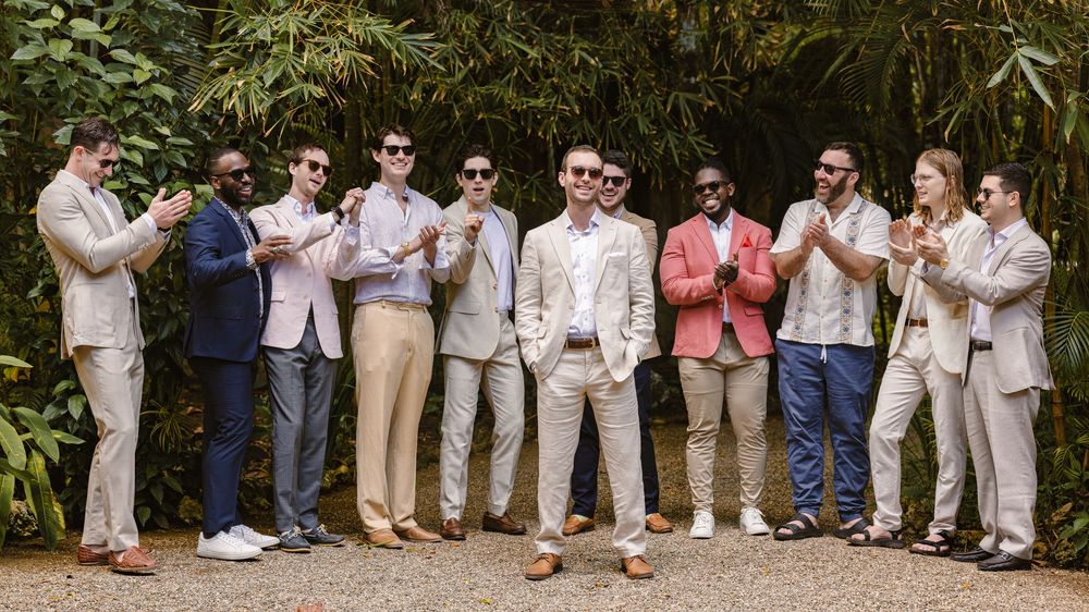 Wedding party members in light colored attire stand in a row against lush greenery at an outdoor venue.