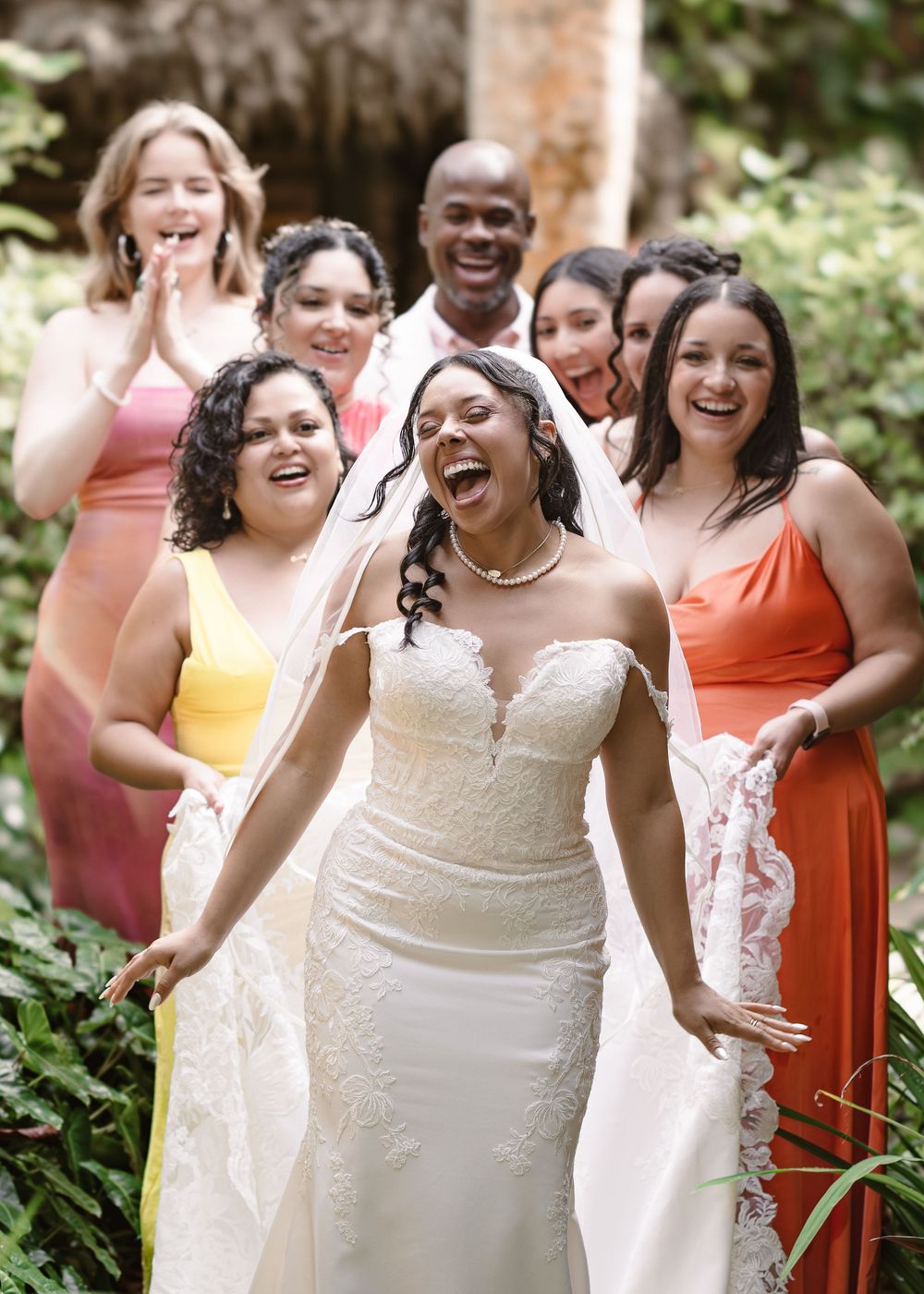 Wedding party in orange and cream dresses laughing joyfully together in an outdoor garden setting.