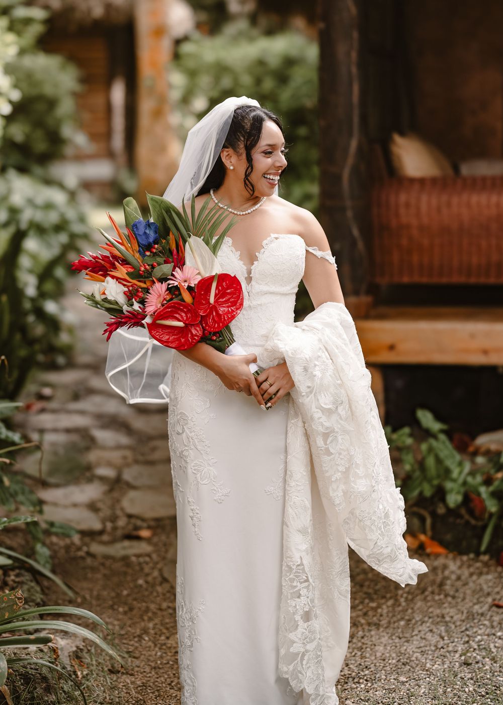 A bride in a lace wedding dress holds a vibrant red and blue tropical bouquet in an outdoor garden setting.