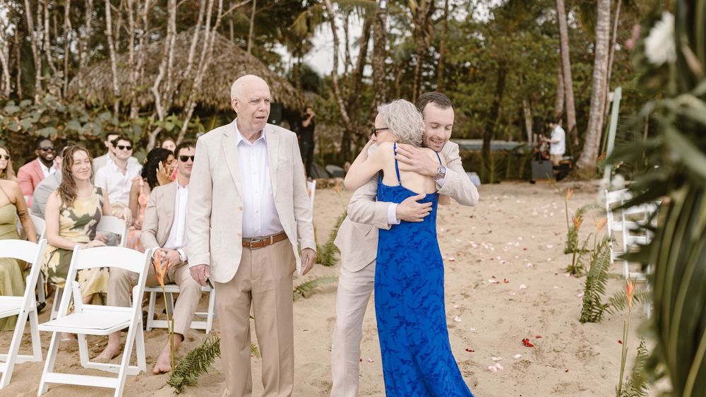 Wedding party stands in a line on a beach with trees in the background during a sunny outdoor ceremony.
