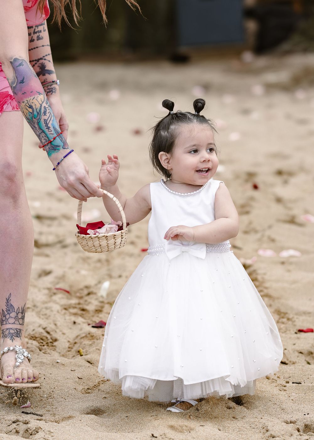 A small child in a white tulle dress stands on sand while someone holds a basket nearby.