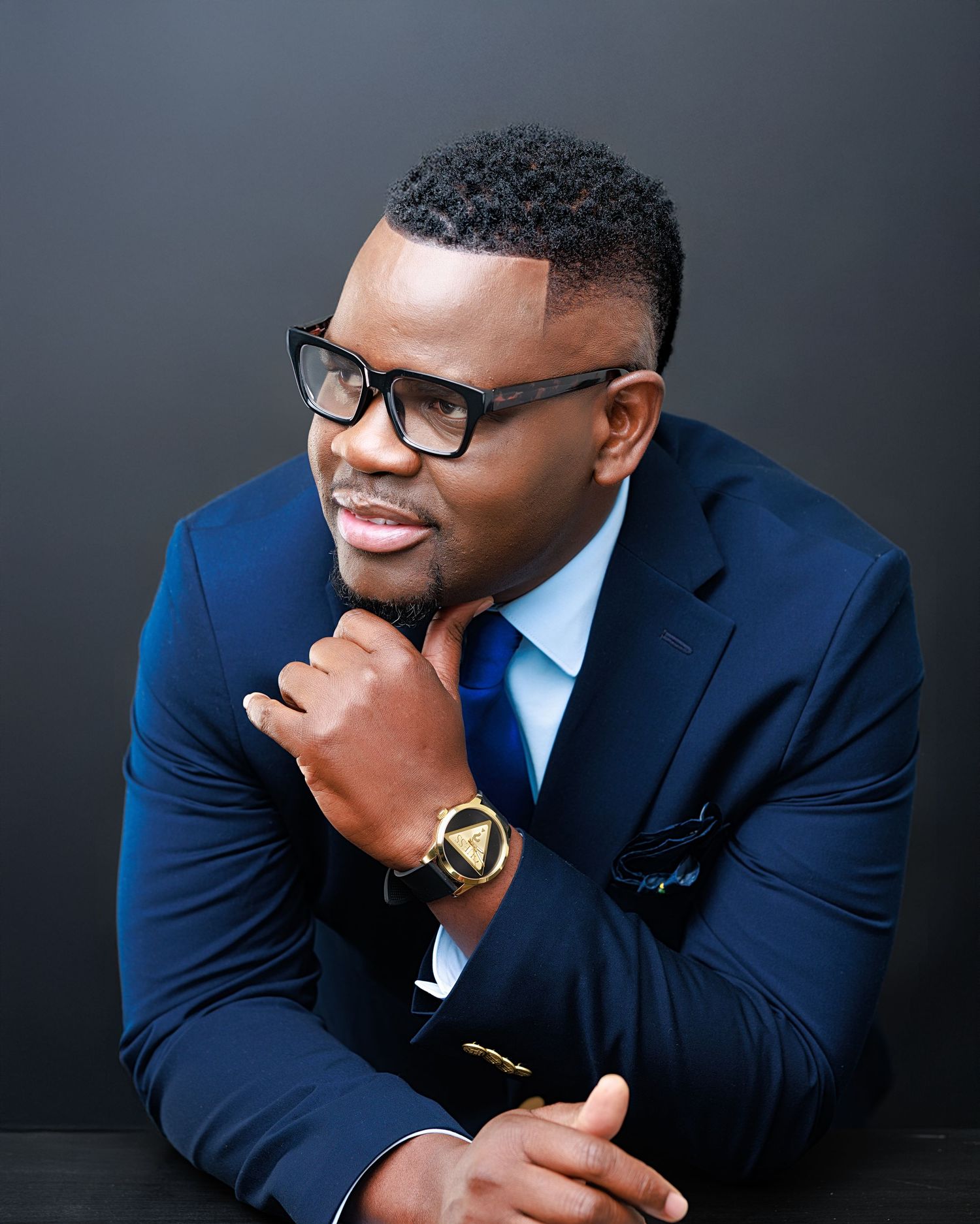 Professional wearing a navy suit and glasses poses thoughtfully against dark background in business portrait series.