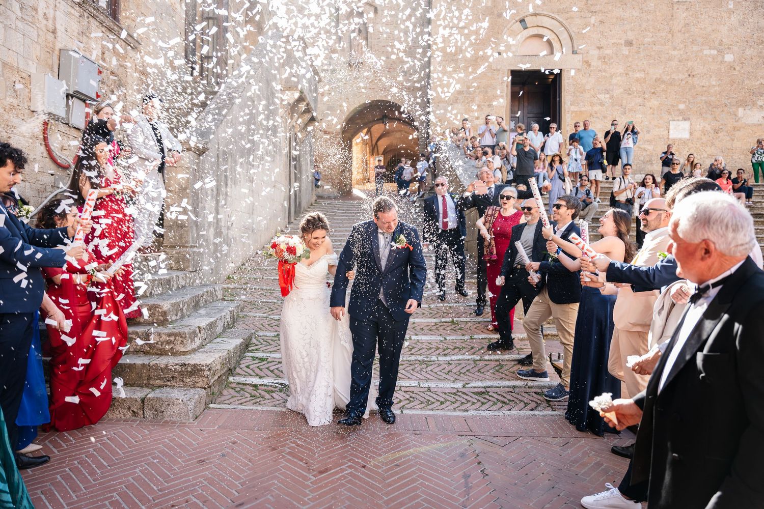 Wedding guests throw rose petals as newlyweds walk down stone steps at historic venue.