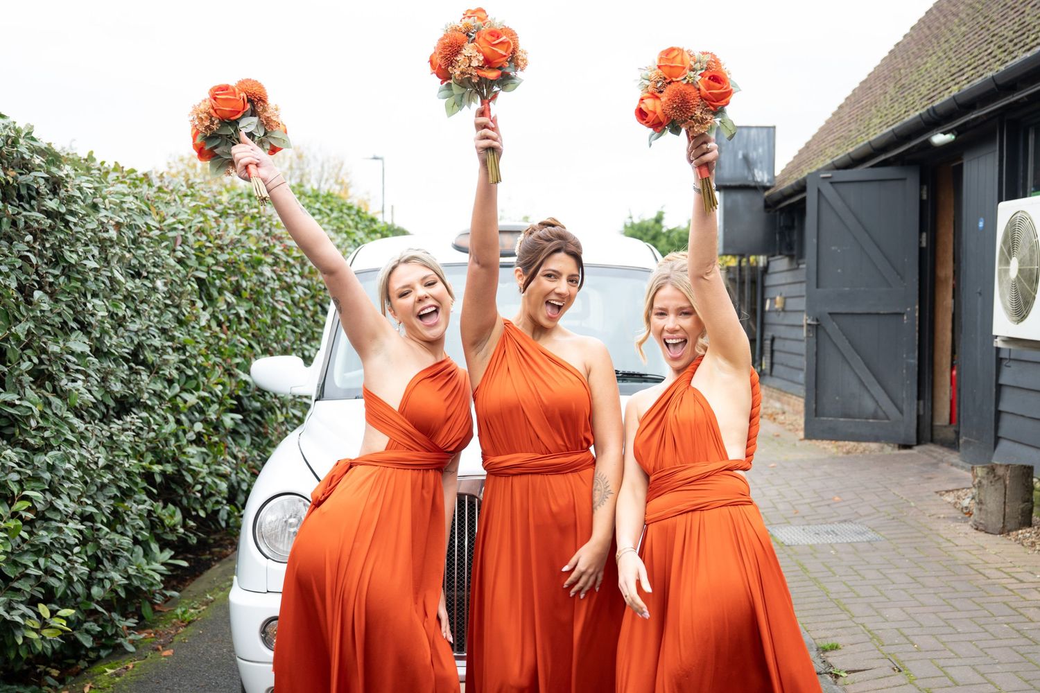 Three people in matching bright orange bridesmaid dresses hold up coral bouquets while celebrating outside.