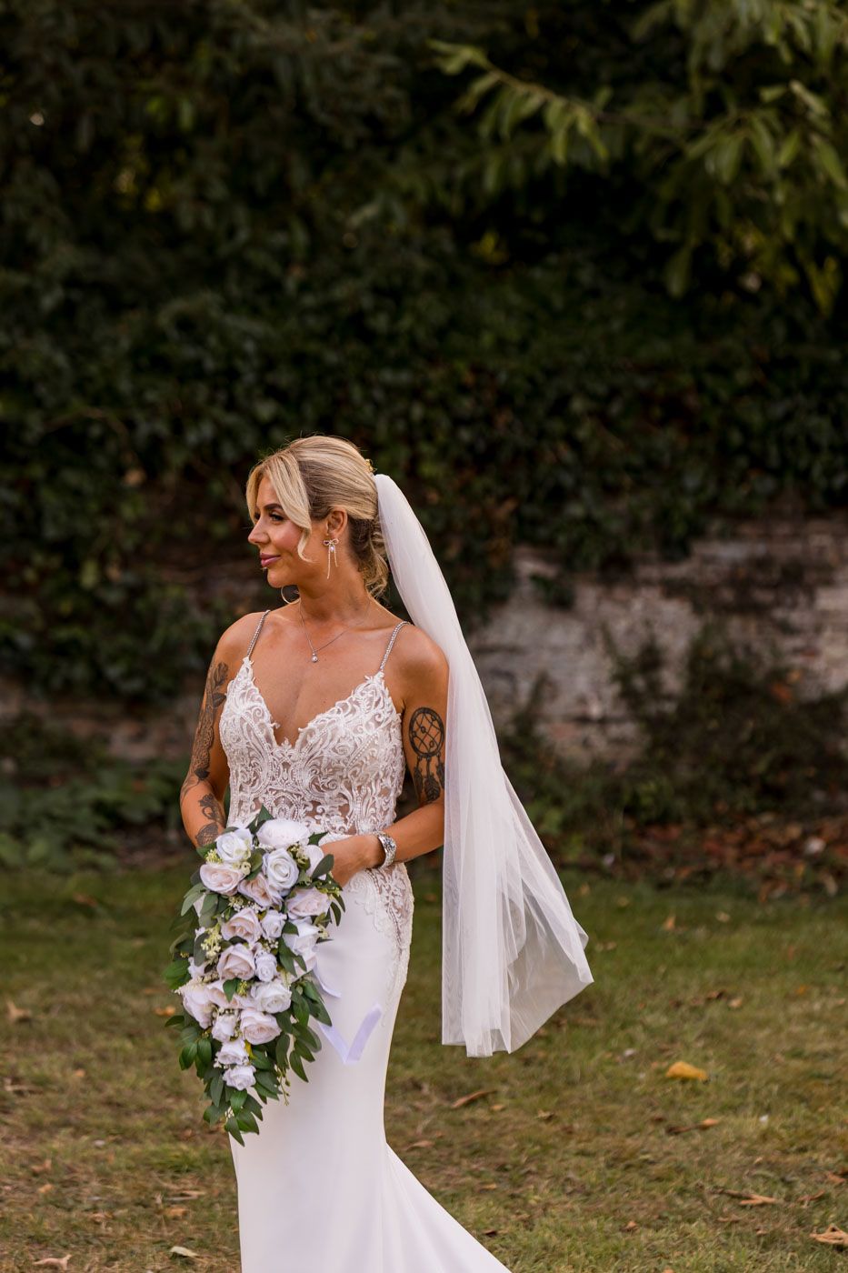 Bride in lace detailed gown holding cascading purple and white bouquet in garden setting.