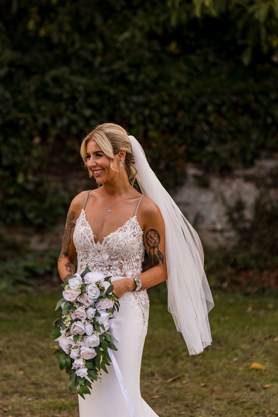 Bride with flowing veil holding purple and white bouquet in outdoor garden setting.