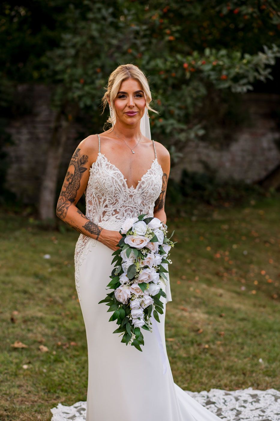 Side view of bride in fitted lace gown with cascading bouquet outdoors.